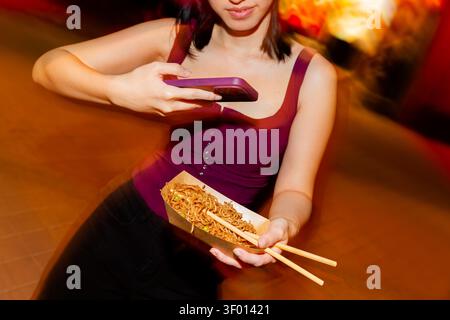 Jeune femme à un marché alimentaire de rue de nuit prenant une photo smartphone de nouilles asiatiques dans un plateau à emporter avec des baguettes, capturant un snac urbain décontracté Banque D'Images
