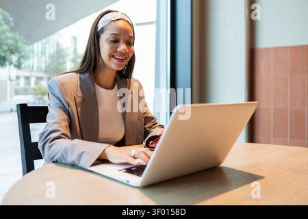 Une femme dans un blazer élégant travaille sur son ordinateur portable dans un café moderne, profitant de l'atmosphère sereine pour se concentrer sur la productivité et la créativité wi Banque D'Images