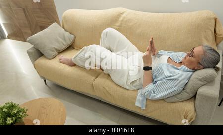 Femme senior allongée sur un canapé beige dans un salon moderne lumineux, défilant sur son smartphone tout en se reposant sur des oreillers, profitant de temps de loisirs tranquilles a Banque D'Images