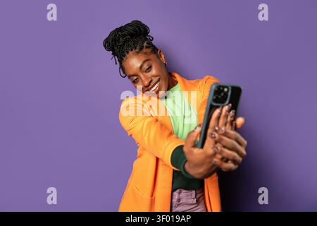 Jeune femme afro dans un cadre de studio prenant un selfie avec un smartphone, souriant avec confiance dans une tenue colorée sur fond violet, moderne l Banque D'Images