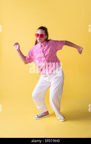 Jeune danseuse posant à mi mouvement dans un studio sur un fond jaune vif, portant une chemise à motifs roses, un pantalon blanc et des lunettes de soleil, expres Banque D'Images