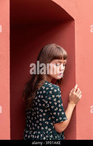 Jeune femme avec de longs cheveux bruns dans une robe florale sombre se tient près d'une arche en stuc rouge, regardant vers le bas dans une lumière douce. Portrait minimal avec ton chaud Banque D'Images
