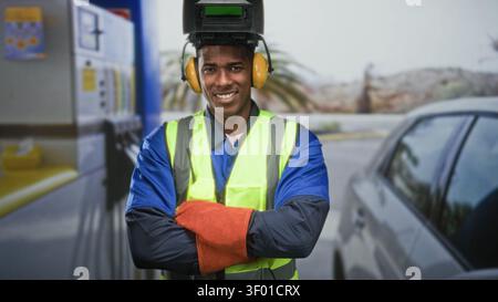 Homme portant un casque de soudure et gilet réfléchissant, les bras croisés et souriant dans la rue près d'une pompe de voiture ; sécurité de confiance. Banque D'Images