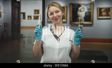 Femme dentiste portant des gants uniformes et bleus tenant des brosses à dents électriques et en bambou dans la galerie du musée souriant ; contraste éducatif. Banque D'Images