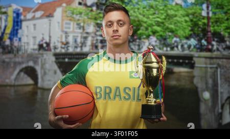 Maillot homme au brésil tient le basket-ball et le trophée, la main droite saisissant le ballon tout en se tenant debout dans la rue ; fierté de la victoire. Banque D'Images