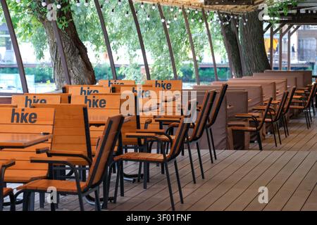 KIEV, UKRAINE, 5 JUILLET 2026, terrasse de restaurant vide en plein air avec des tables et des chaises en bois préparées pour les visiteurs un jour d'été le long de la rivière. Banque D'Images