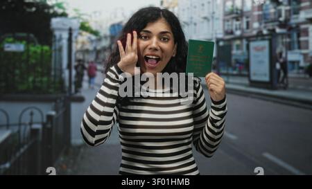 Jeune femme brune tient le passeport vert dans la main droite et sourit tout en appelant dans la rue de la ville ; fierté de la liberté de voyager. Banque D'Images