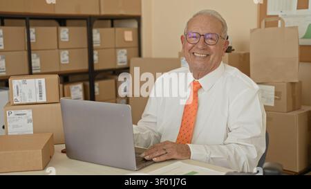Homme souriant avec les mains sur l'ordinateur portable tapant au bureau parmi les colis empilés et les boîtes dans un bâtiment de salle d'expédition ; confiance entrepreneuriale. Banque D'Images