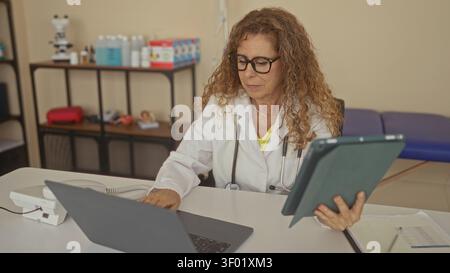 Femme médecin tenant la tablette et tapant sur ordinateur portable à la clinique, portant un manteau blanc et stéthoscope tout en examinant les dossiers médicaux ; soins ciblés. Banque D'Images