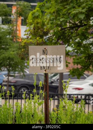 Un panneau anti-intrusion BNSF altéré sur un poteau en métal rouillé derrière des plantes vertes avec des voitures floues et des arbres en arrière-plan Banque D'Images