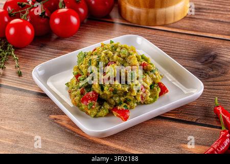 Guacamole frais avec des morceaux de poivron rouge sur une assiette carrée blanche. Fond de table en bois rustique avec tomates cerises piments rouges Banque D'Images
