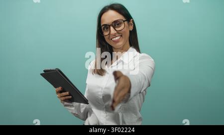 Jeune femme hispanique atteint la main pour une poignée de main tout en tenant la tablette et en souriant sur fond de studio sarcelle ; professionnel accueillant. Banque D'Images
