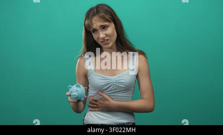 Jeune femme tenant une tirelire bleue et serrant le ventre avec une main dans le studio sarcelle ; inquiétude financière de la dette. Banque D'Images
