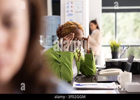 Femme afro-américaine se penche vers l'avant au bureau avec les yeux fermés, traitant de maux de tête de tension battant. Gestionnaire féminin ressent la fatigue et la tension émotionnelle des délais constants dans le bureau de l'entreprise. Banque D'Images