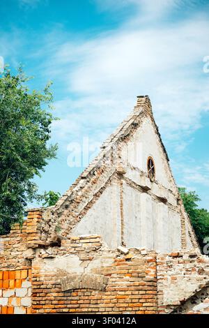 Bâtiment en ruine de briques altérées avec pignon triangulaire abrupt et fenêtre ronde placé contre un ciel bleu vif, entouré d'arbres et de maçonnerie en ruine qui s'effondre Banque D'Images