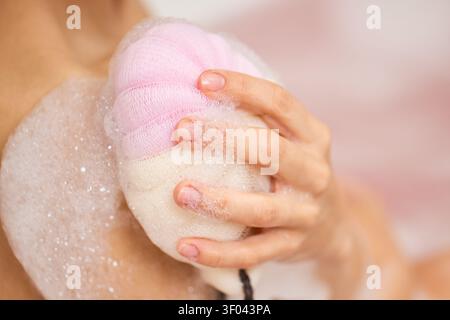 Main de femme tenant une éponge de bain rose, frottant sa peau et créant d'abondantes bulles de savon blanches lors d'un bain relaxant pour l'hygiène Banque D'Images