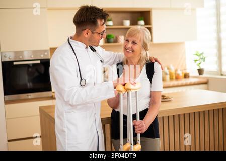 Un professionnel de la santé encourage une femme âgée à utiliser des béquilles d'avant-bras lors d'une visite à domicile. Banque D'Images