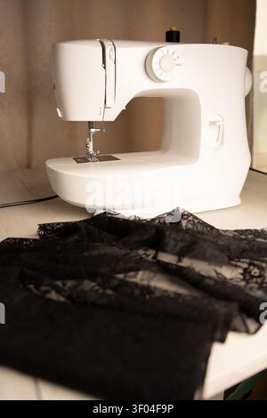 gros plan légèrement éclairé d'une machine à coudre blanche sur une table en bois, aiguille posée au-dessus de délicate dentelle noire, toile de fond beige chaude évoquant un intime, Banque D'Images