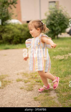 jeune fille joyeuse dans une robe à rayures pastel avec des queues de cochon et des sandales roses en cours d'exécution et tourbillonnant sur un chemin de gravier à côté des buissons verts et de l'appartement b. Banque D'Images