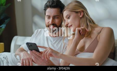 Famille optimiste couple caucasien discutant de l'application à la recherche dans le téléphone portable dans le lit homme avec femme discuter de la décision sur les achats en ligne e Banque D'Images