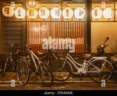 Rangée de lanternes en papier blanc illuminées suspendues au-dessus de vélos stationnés devant une vitrine de restaurant la nuit, Kyoto, Japon Banque D'Images