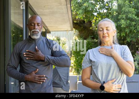 Couple diversifié pratiquant la respiration rythmée sur le patio en vêtements athlétiques avec smartwatch. Respiration, pleine conscience, bien-être, terrasse, extérieur, ensoleillé, nature Banque D'Images