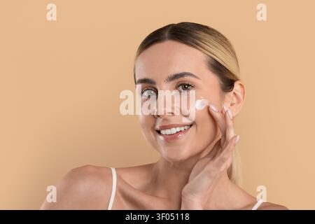 Jeune belle femme souriante appliquant une crème de soin sur sa joue Banque D'Images