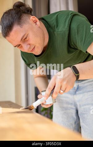 Homme d'âge moyen applique la teinture de bois avec un pinceau sur une planche de bois tout en travaillant sur un projet de mobilier DIY sur un balcon. Concept de maison rénovati Banque D'Images