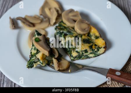 Morceaux de frittata aux épinards servis avec des champignons tranchés sur une assiette blanche, offrant un repas sain et riche en protéines Banque D'Images