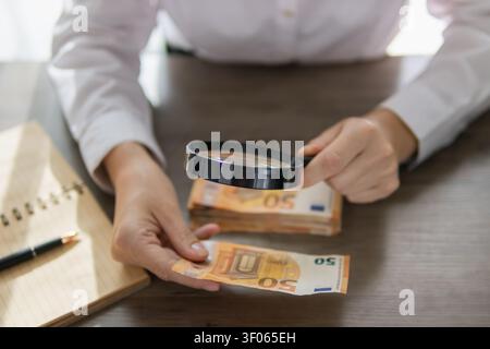 Personne tenant une loupe, inspectant de près un billet de 50 euros sur un bureau en bois avec une pile d'argent et un carnet, en se concentrant sur les aspects financiers Banque D'Images