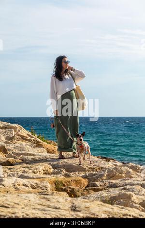 Femme enceinte promenant son terrier sans poil américain sur une plage rocheuse au bord de la mer, profitant d'une journée ensoleillée à l'extérieur avec laisse et sac fourre-tout, vie de vacances Banque D'Images