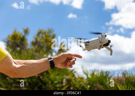 Gros plan de la main d'une personne atteignant un petit drone quadcopter planant à l'extérieur par une journée brillante, avec un ciel bleu, des nuages et des arbres verts dans le Banque D'Images