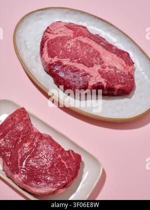 Deux steaks de bœuf frais crus sont affichés sur d'élégantes assiettes blanches sur un fond rose doux. La viande rouge éclatante met en valeur le marbrage, idéal pour emporter Banque D'Images