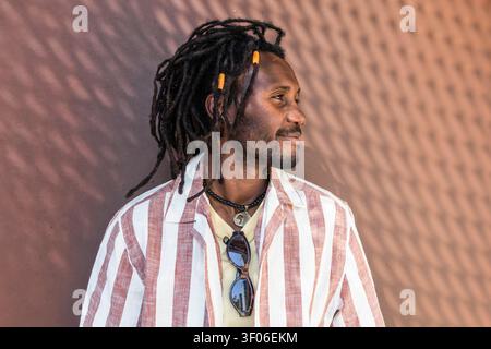 Portrait profil de côté d'un homme cubain avec des dreadlocks et collier de perles, portant une chemise rayée et des lunettes de soleil, debout près d'un mur texturé au chaud Banque D'Images