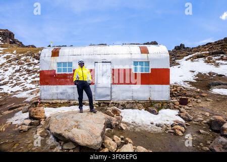 Randonneur vêtu d'un équipement lumineux se tient devant un abri de montagne rustique entouré d'un terrain rocheux et enneigé, mettant en vedette un backpa en plein air aventureux Banque D'Images