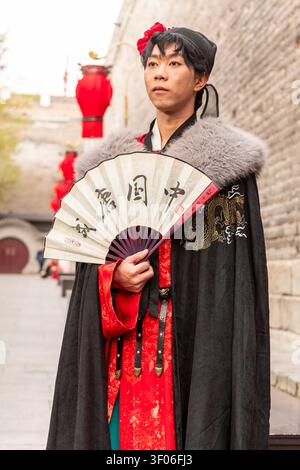 Jeune homme portant le hanfu chinois traditionnel et la cape tient un éventail pliant tout en se tenant debout le long de l'ancien mur de la ville de Xi'an en Chine, avec des lanternes rouges Banque D'Images