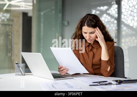 Jeune travailleuse subissant des maux de tête et du stress en examinant des documents financiers, se sentant submergée et inquiète face aux problèmes commerciaux et à la paperasse exigeante au bureau Banque D'Images