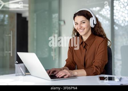 Jeune femme d'affaires souriant tout en tapant sur son ordinateur portable à un bureau, portant des écouteurs blancs et fournissant un support client à distance Banque D'Images