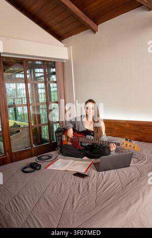 Jeune musicienne pratiquant la basse guitariste tout en suivant un cours en ligne sur un ordinateur portable dans une chambre confortable. Banque D'Images