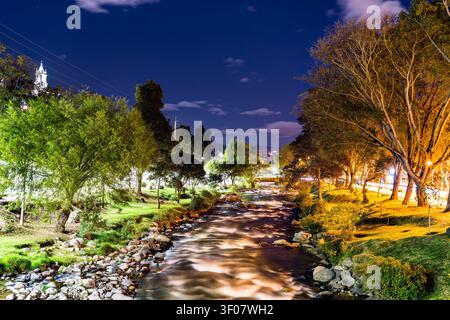 Belle vue nocturne longue exposition sur la rivière Tomebamba qui traverse Cuenca, Équateur, entourée d'arbres luxuriants et de lumières de la ville. Banque D'Images