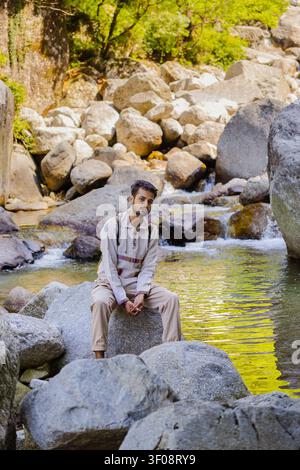 Un jeune homme avec un sac à dos est assis sur un gros rocher à côté d'un ruisseau de montagne qui coule dans un paysage rocheux. Banque D'Images