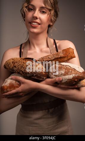Boulangère souriante portant un tablier et tenant une variété de pains au levain artisanaux fraîchement cuits Banque D'Images