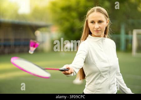 Joueuse de badminton confiante frappant un volant en plein vol sur un terrain vert ensoleillé. Sport d'été, précision et action à l'heure dorée. Banque D'Images