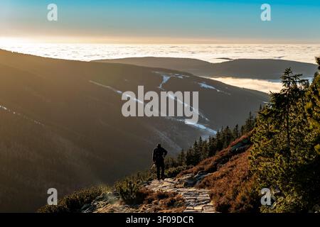 Silhouette d'un randonneur solitaire descendant un sentier de pierre montagneux au coucher du soleil doré surplombant une vaste mer de nuages remplissant les vallées en contrebas Banque D'Images