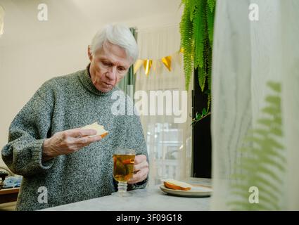 Un senior de plus de 70 ans déguste un petit-déjeuner composé de thé et de sandwich au beurre, mettant l'accent sur une alimentation équilibrée. L'image promeut des habitudes alimentaires saines dans la vieillesse. Grand-père attentif à son régime. Banque D'Images