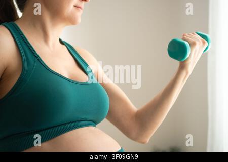 Une femme enceinte tient un haltère dans une pose détendue et décontractée contre un mur blanc à l'intérieur. La mère et athlète montre une confiance à l'aise et une force calme et naturelle lors d'un entraînement à domicile. Banque D'Images