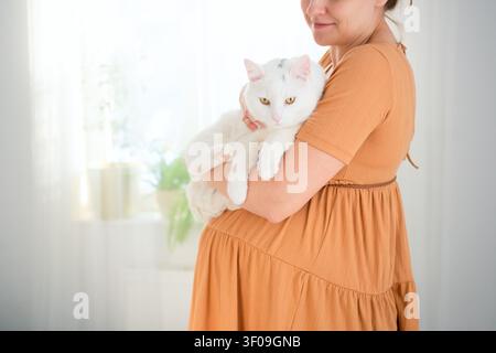 Une femme enceinte tient un chat blanc dans ses bras dans une pièce lumineuse. La future mère câlinant son animal de compagnie présente un moment calme et paisible avec une conscience de la prévention des maladies et de la protection. Banque D'Images