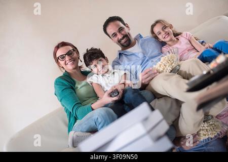 Une famille moderne arabe du moyen-Orient composée de quatre personnes se détend sur un canapé, souriant et partageant du pop-corn tout en regardant un film à la maison, transmettant chaleur, convivialité, confort décontracté et joyeux liens familiaux. Banque D'Images