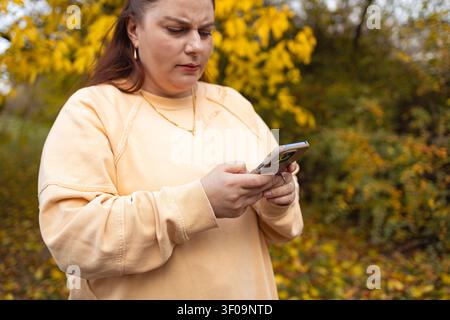 Femme envoyant un SMS sur son téléphone dans une scène automnale en plein air avec des arbres et du feuillage, gros plan, légèrement bas, mise au point, sweat-shirt beige, coque en plastique Banque D'Images