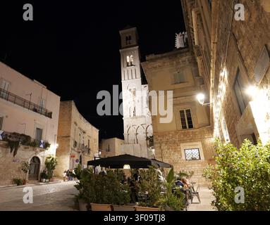 Place de la ville de Giovinazzo la nuit, avec le clocher roman de la cathédrale Santa Maria Assunta se dressant fièrement, des gens profitant d’un dîner en plein air. Italie Banque D'Images